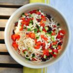 Noodles with Greek Salad ingredients in a bowl on a yellow striped dish towel