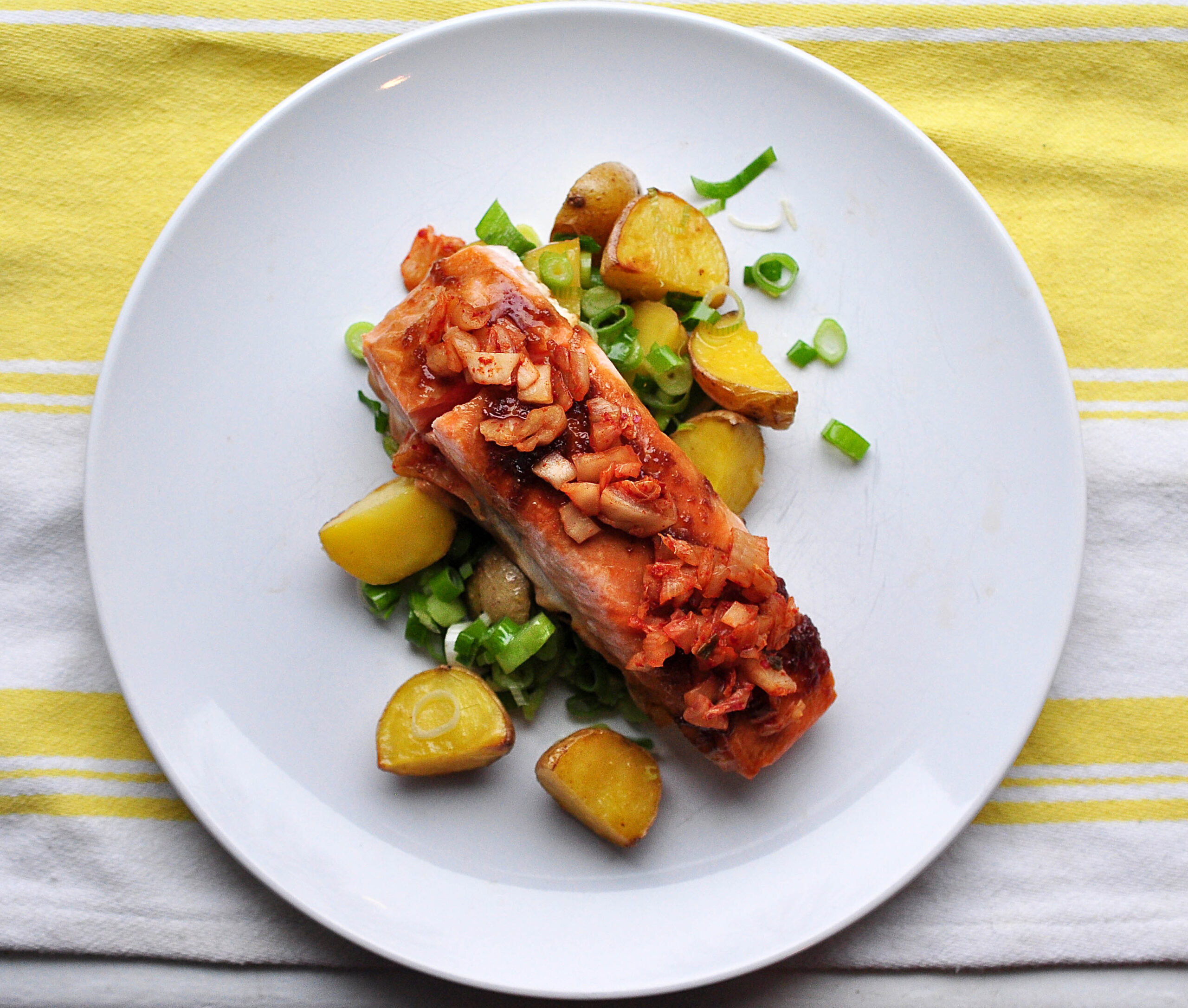 white plate with pile of roasted potatoes and scallions and a fillet of salmon glazed with soy and garlic and topped with kimchi