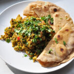 white plate with pile of very yellow scrambled eggs and vegetables with cilantro on top and four pieces of roti