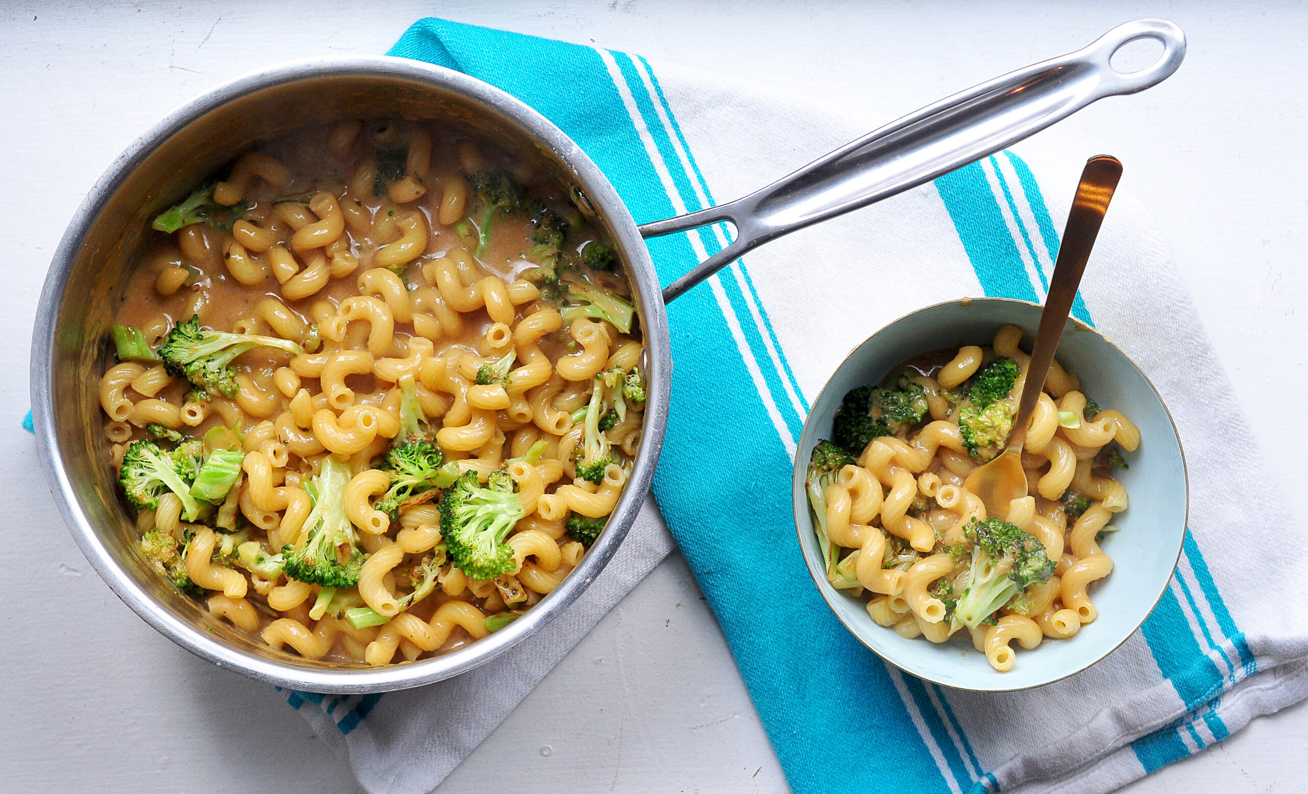 welsh rarebit macaroni and cheese with broccoli in a pot on a teal striped tea towel and in a bowl with a fork