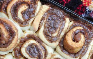 Snail-like rolls of golden brown dough bubbling over with a dark brown filling of sugary goo arranged in a baking tray with a red and green and black checked blanket nestled against the baking tray. Set at an angle.